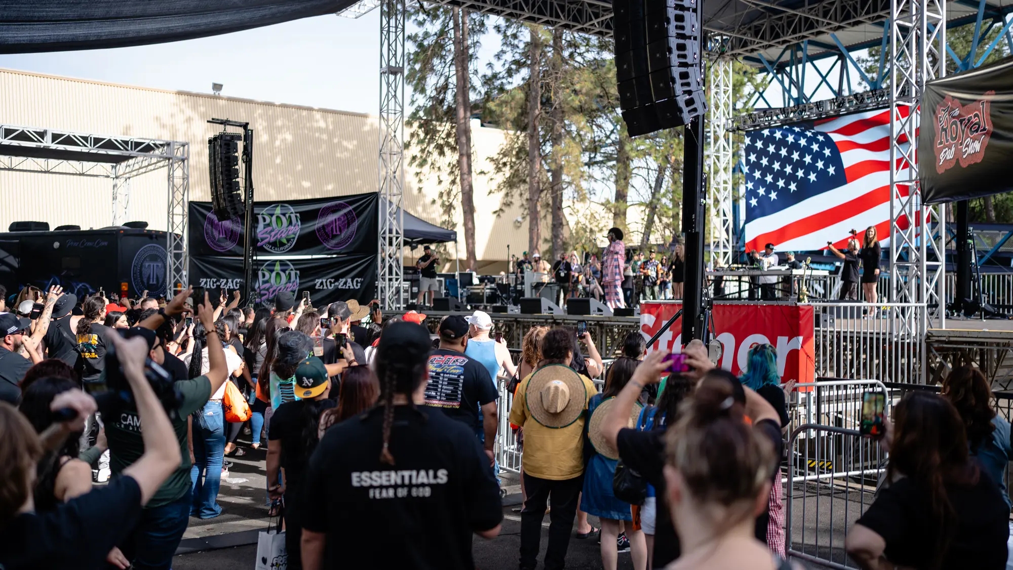 A large outdoor crowd holding up phones and cameras toward a festival stage with an American flag screen behind the performers.