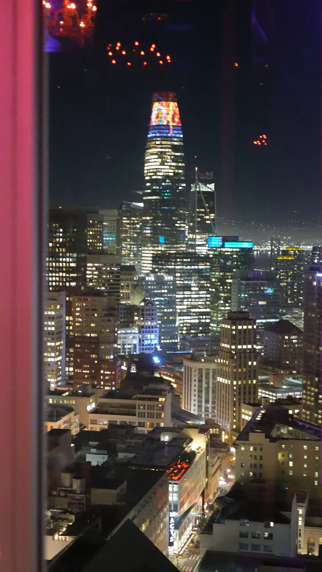 San Francisco's skyline at night with a lit skyscraper rising above downtown buildings, seen through a window.