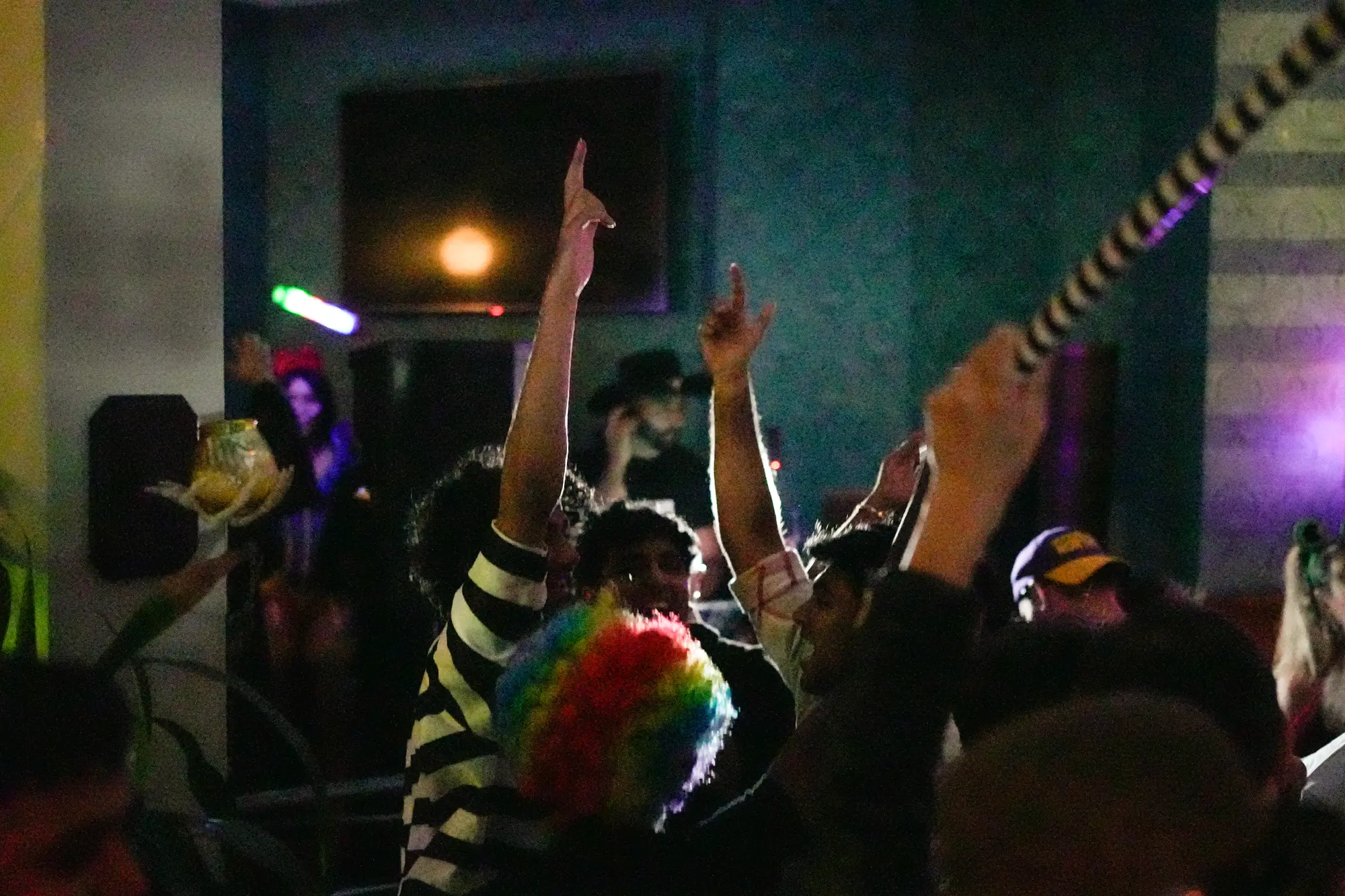 Silhouetted guests, one in a rainbow wig, raise their arms toward the stage light in a dim room.