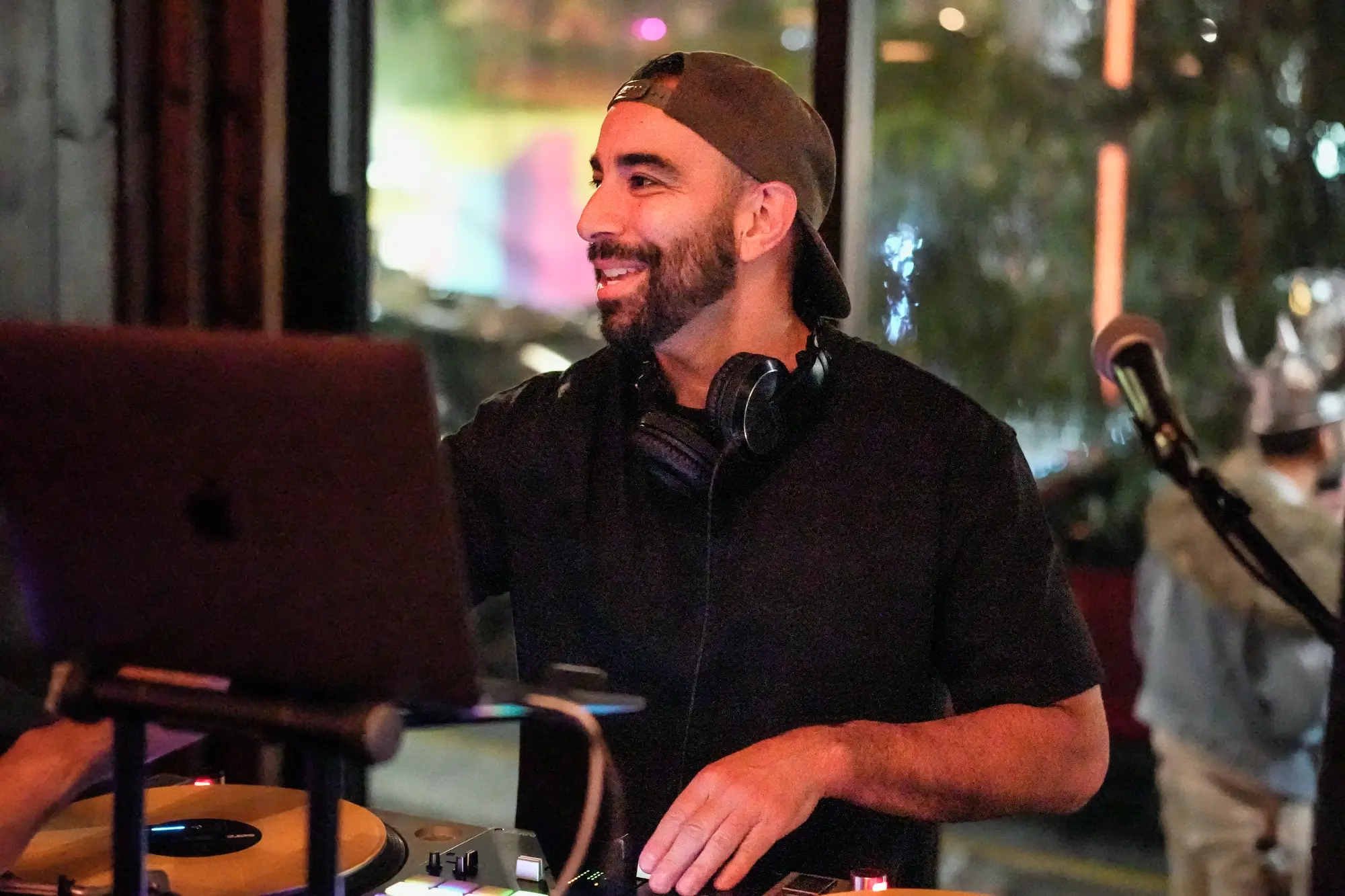 DJ Mojo smiling over his laptop with a hand on the mixer, headphones around his neck.