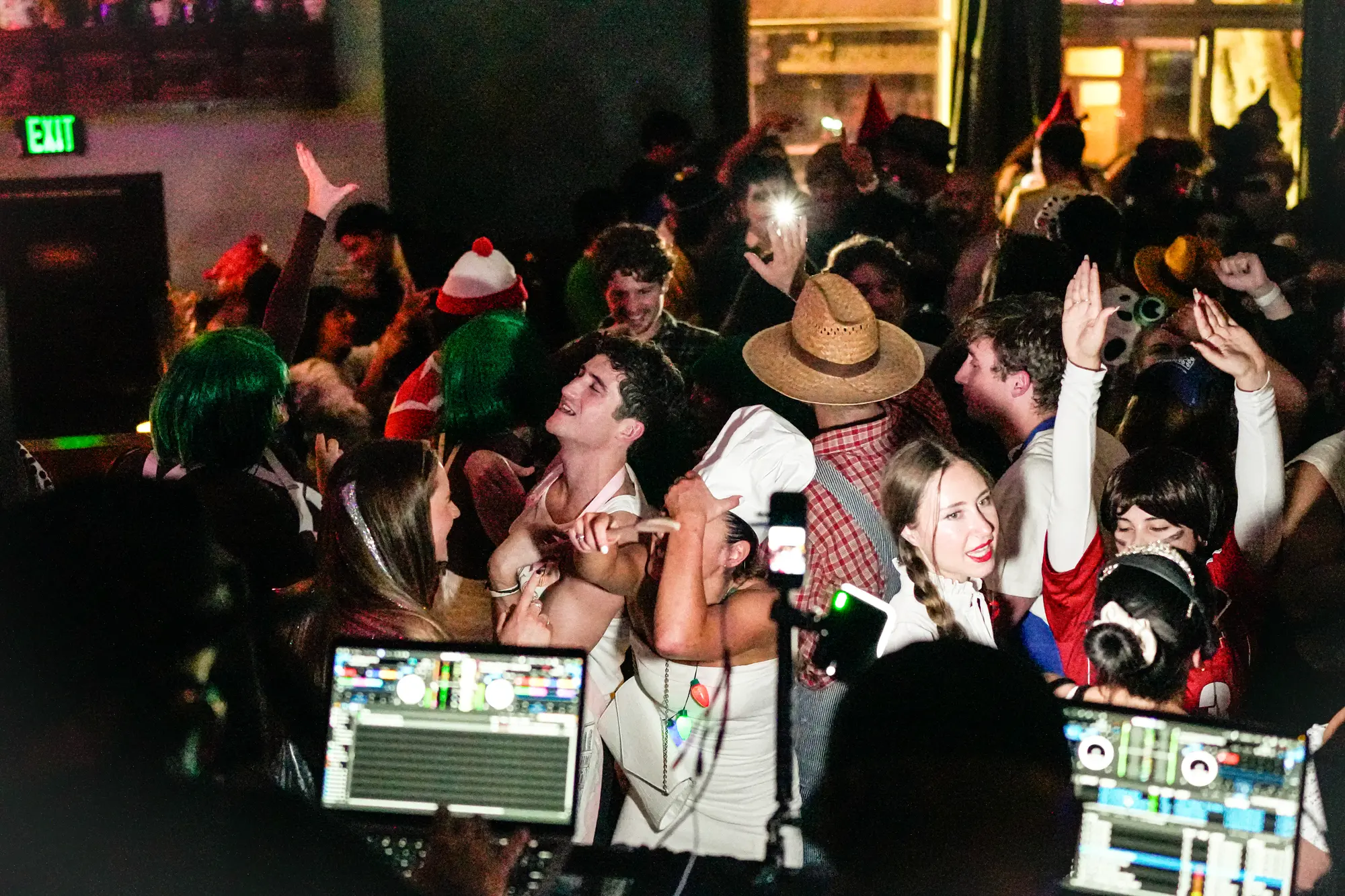 A packed dance floor of costumed guests with hands in the air, with the DJ's laptops glowing at the bottom of the frame.