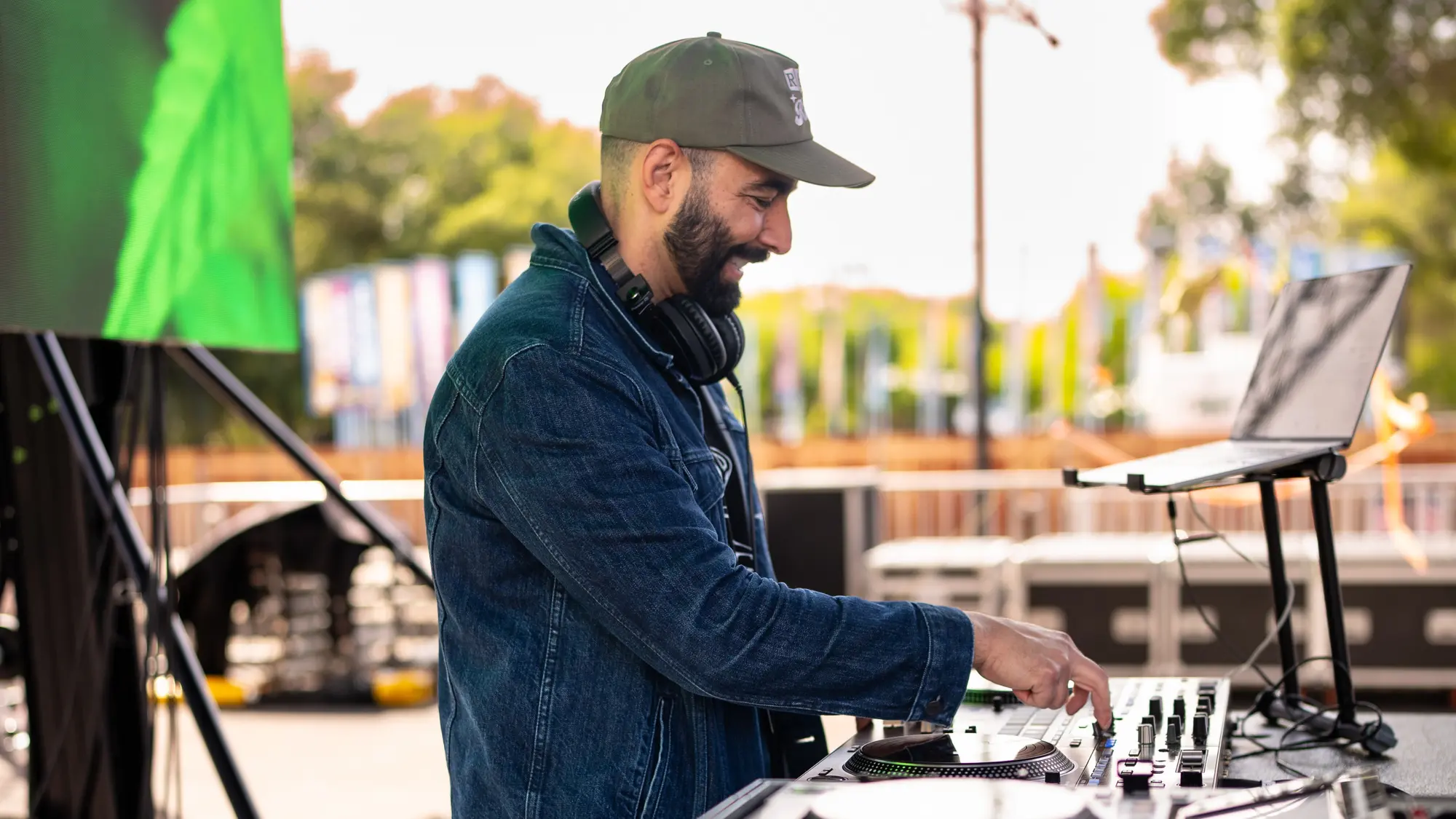 DJ Mojo in a cap and denim jacket, smiling as he mixes on an outdoor stage with a laptop beside him.